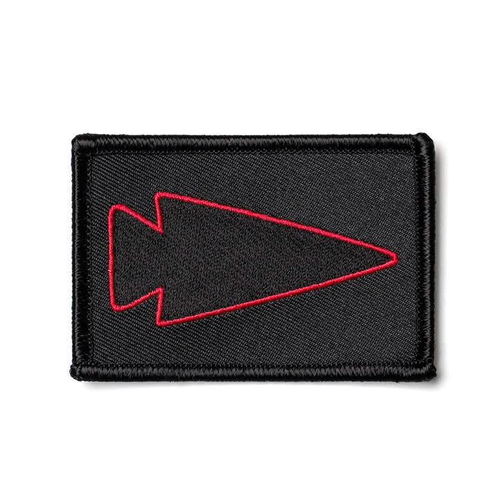 The Patch - Spearhead Outline by GORUCK is a black rectangular embroidered patch that showcases a red outline of an arrowhead pointing to the right at its center. The neatly stitched edges enhance the sleek design, complemented by the distinctive touch of the GORUCK logo.
