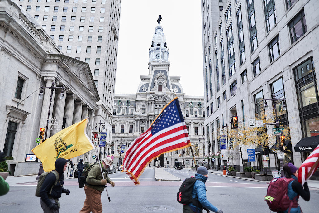 People march with American and yellow flags near Philadelphia City Hall on a city street.