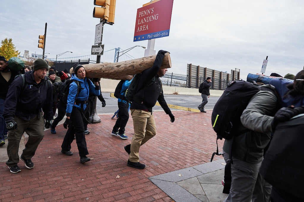 A group of people walk on a city sidewalk carrying a large log together near a Penn’s Landing sign.
