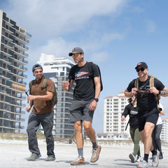 Three people carrying Mackall 45 Fog backpacks walk on a beach near tall buildings, smiling and enjoying the sunny weather.