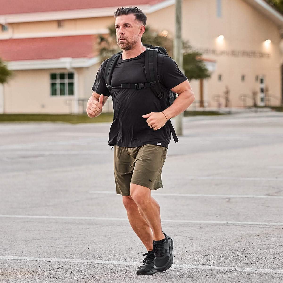 A person in a black t-shirt and shorts, sporting the GORUCK Men's Rough Runner - Blackout running shoes, jogs across a parking lot with a black backpack. In the background, a building with a red roof and beige walls stands under the sun.