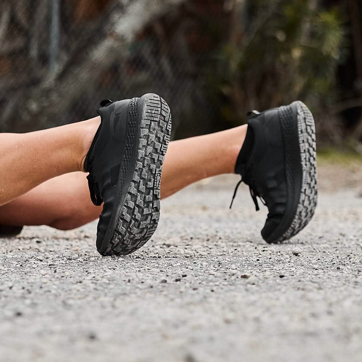 A person lying on a gravel path, sporting GORUCK's Men's Rough Runner - Blackout athletic shoes with rugged soles. The focus is on the shoes, while the background reveals blurred greenery and a fence.