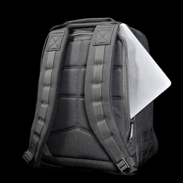 A black Radio Ruck USA Throwback backpack with padded straps, partially unzipped to show a laptop securely stored in its bombproof compartment against a black background.