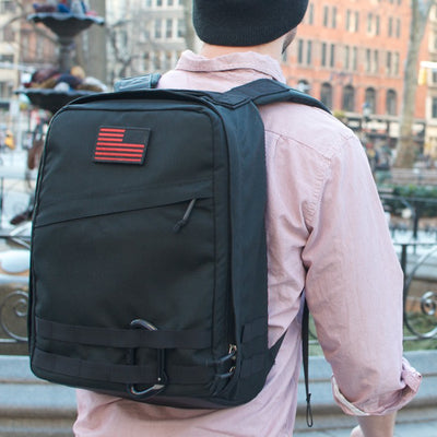 A person stands outdoors in an urban area wearing a black Radio Ruck USA Throwback backpack with a red-striped patch and bombproof laptop compartment.