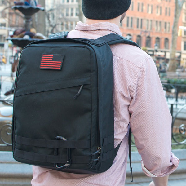 A person stands outdoors in an urban area wearing a black Radio Ruck USA Throwback backpack with a red-striped patch and bombproof laptop compartment.