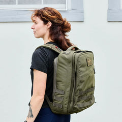 A person with brown hair tied back is standing sideways, wearing a black T-shirt and carrying the GORUCK GR2 - Cordura in green. They are outdoors in front of a light-colored wall with a window above, embodying the spirit of the Special Forces lifestyle.