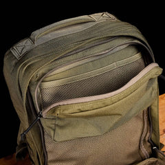 A close-up of a green GR2 - Cordura by GORUCK highlights its numerous zippered compartments. The ruck is open, displaying mesh-lined pockets, set against a dark background on a wooden surface, evoking an aura of Special Forces efficiency.