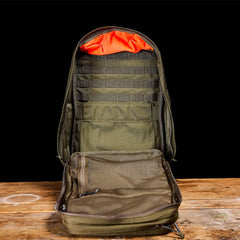 A GORUCK GR2 tactical backpack, inspired by Special Forces quality and featuring an open design with a bright orange lining at the top, multiple inner compartments, and mesh pockets, is displayed on a wooden surface against a black background.