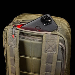 The GR2 - Cordura by GORUCK, a black smartphone rests in the partially open exterior pocket of a green, textured ruck against a black background; red accents highlight the zipper pull and inside the pocket, reminiscent of Special Forces precision.