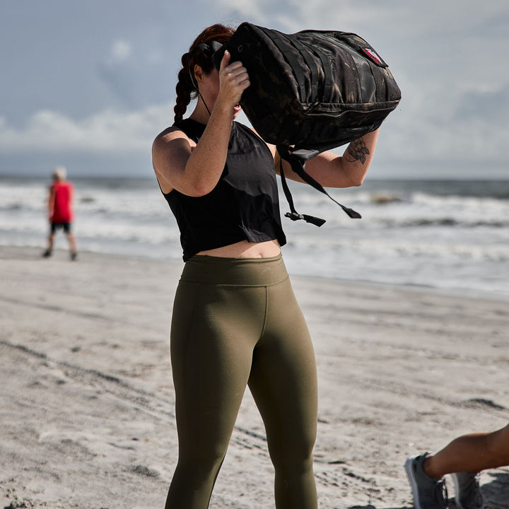 On the beach with a stunning ocean backdrop, a woman lifts a sandbag wearing GORUCK Women’s Training Leggings - ToughFlex, designed from durable ToughFlex Fabric for squat-proof performance.