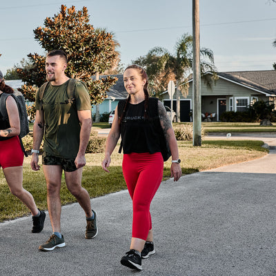Three individuals walk down a suburban street, backpacks in tow, dressed in athletic gear. The women wear GORUCK Women's Cropped Training Leggings made from ToughFlex fabric, providing squat-proof comfort for their outdoor adventure.