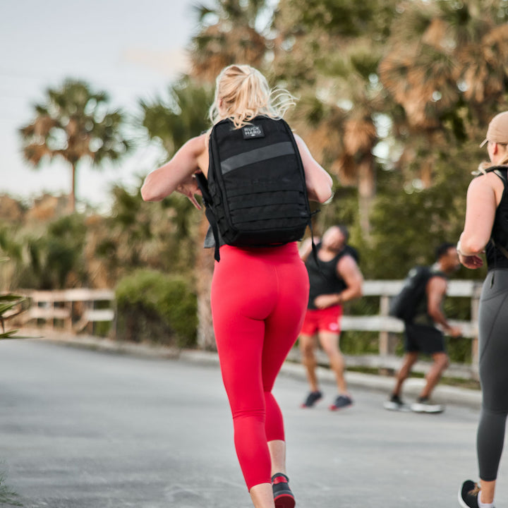 Wearing GORUCK Women’s Training Leggings - ToughFlex, ideal for being squat proof, a person is jogging on a path. Their black backpack bounces gently with each stride, surrounded by trees and the lively presence of people in the background.