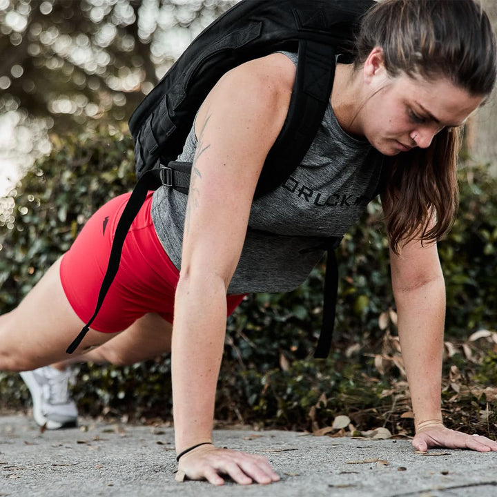 A person doing a push-up outside is wearing GORUCK's Women's Squat Shorts in vibrant red, designed for full freedom of movement, along with a gray top and a backpack. The ToughFlex fabric offers durability and comfort, while the no-ride technology ensures the shorts stay perfectly in place during the workout.
