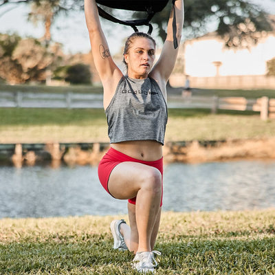 Exercising outdoors near a tranquil pond and trees, the person lunges with weights overhead, wearing the Women’s Squat Shorts - ToughFlex by GORUCK, designed with stay-put technology for ultimate comfort and flexibility.