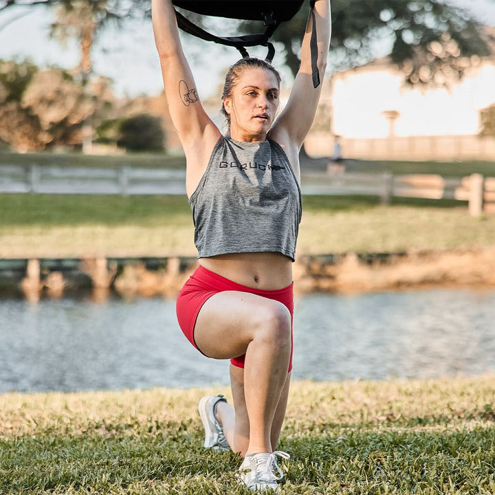 Exercising outdoors near a tranquil pond and trees, the person lunges with weights overhead, wearing the Women’s Squat Shorts - ToughFlex by GORUCK, designed with stay-put technology for ultimate comfort and flexibility.