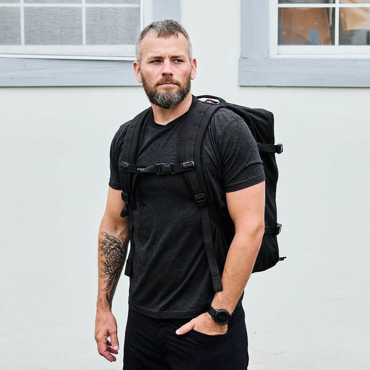 A man with short hair and a beard stands against a light-colored wall. He wears a dark t-shirt, black backpack, and a watch. A tattoo is visible on his left arm as he confidently carries his sturdy GR3 - Ripstop ROBIC® by GORUCK.
