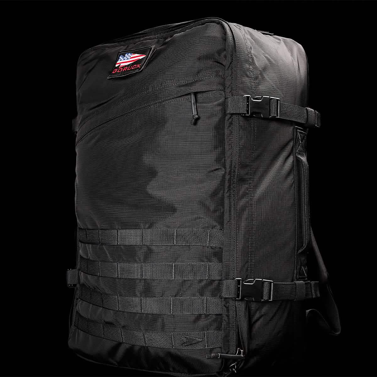 The GR3 - Ripstop ROBIC® by GORUCK is a large black tactical carry-on rucksack made from durable Robic nylon. It boasts multiple straps and buckles, and features a small patch with the American flag and "GORUCK" lettering on the upper part set against a solid black background.