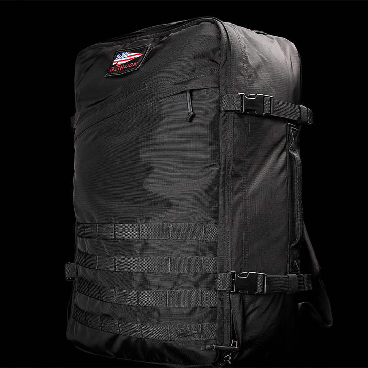 The GR3 - Ripstop ROBIC® by GORUCK is a large black tactical carry-on rucksack made from durable Robic nylon. It boasts multiple straps and buckles, and features a small patch with the American flag and "GORUCK" lettering on the upper part set against a solid black background.