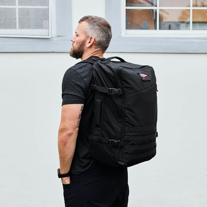 A man with a beard, dressed in a black T-shirt and wearing a smartwatch, carries a GORUCK GR3 - Ripstop ROBIC® rucksack. He stands in profile against a light-colored wall with a window in the background.