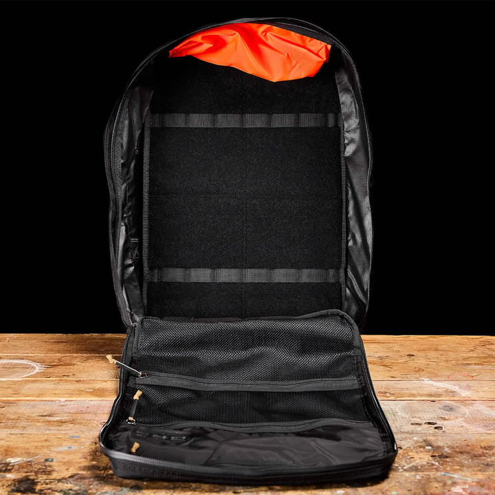 Open the black GR3 - Ripstop ROBIC® backpack by GORUCK against a black background. This carry-on rucksack reveals its spacious interior with an orange lining and a zippered mesh compartment, all crafted from durable Robic nylon. It rests elegantly on a wooden surface.