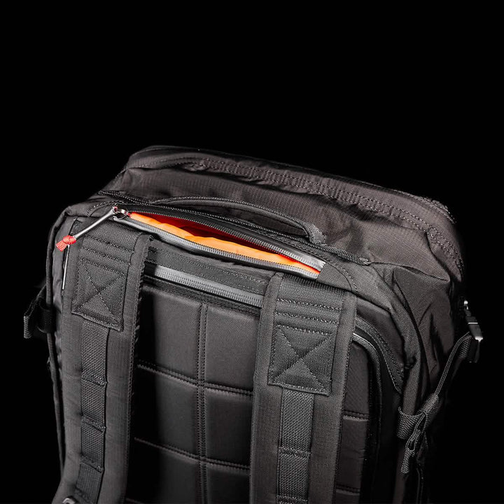 A close-up of a black GORUCK GR3 - Ripstop ROBIC® backpack set against a dark backdrop. The image highlights the top section, featuring padded straps, reinforced stitching in durable Ripstop ROBIC® material, and an orange-lined zipper pocket that is partially open.