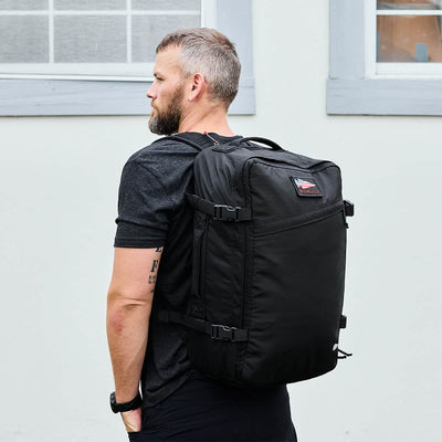 A person with short hair and a beard is wearing a black T-shirt and carrying the large GR3 - Ripstop ROBIC® backpack by GORUCK, designed like a sleek carry-on rucksack, while standing sideways against a light-colored wall with a window.