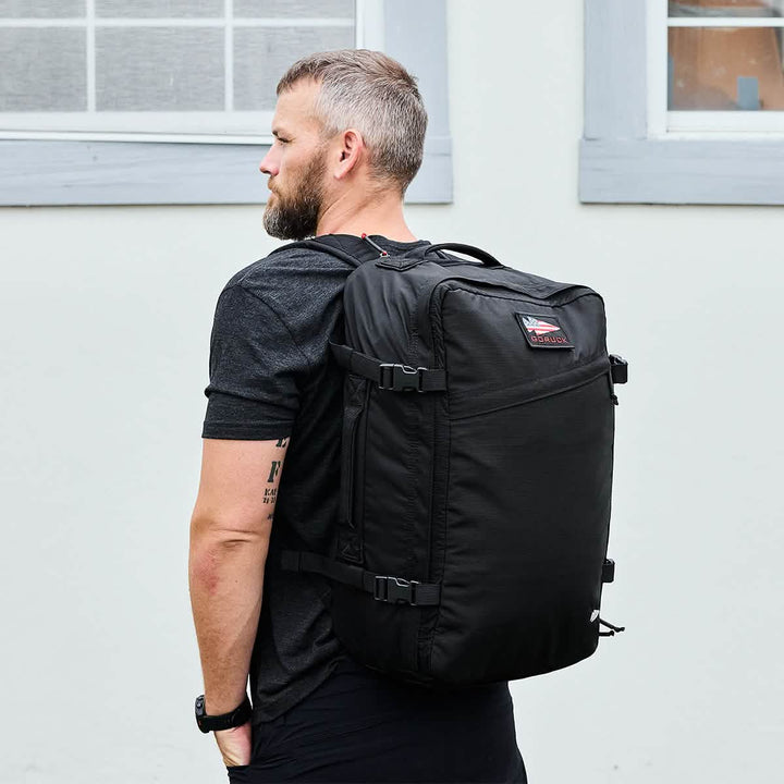 A person with short hair and a beard is wearing a black T-shirt and carrying the large GR3 - Ripstop ROBIC® backpack by GORUCK, designed like a sleek carry-on rucksack, while standing sideways against a light-colored wall with a window.