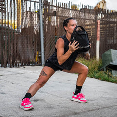 Holding a black weighted bag, a person performs a lateral lunge outdoors. They are sporting GORUCK's Women's Rough Runner - Hot Pink lightweight running shoes, paired with a black tank top and shorts. The vibrant pink of their shoes stands out against the fence and surrounding greenery.