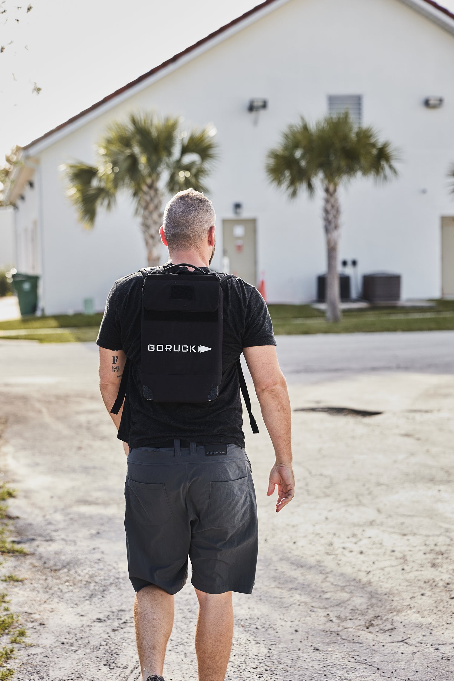 A man with short hair wears the RPC Long + 30LB (Long) Plate Bundle as he walks toward a white building with palm trees, likely doing weighted training.
