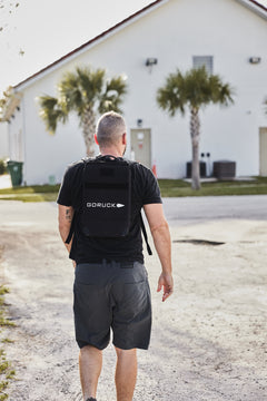 A man with short hair wears the RPC Long + 30LB (Long) Plate Bundle as he walks toward a white building with palm trees, likely doing weighted training.