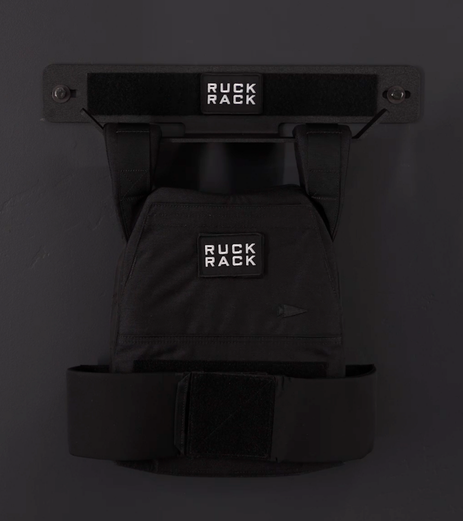 A black Ruck Rack® Delta | Heavy-Duty Wall Mount Storage for Rucks, Weight Vests & Tactical Gear holds a black rucksack labeled “Ruck Rack,” offering a sleek and practical wall-mounted solution for organizing plate carriers and other tactical gear.