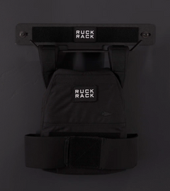 A black Ruck Rack® Delta | Heavy-Duty Wall Mount Storage for Rucks, Weight Vests & Tactical Gear holds a black rucksack labeled “Ruck Rack,” offering a sleek and practical wall-mounted solution for organizing plate carriers and other tactical gear.