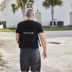 Man wearing a black Ruck Plate Carrier 3.0 made of durable ballistic nylon, walking on a sunny street with palm trees and buildings in the background.