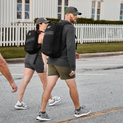 Two individuals sporting GORUCK gear enjoy rucking down a street with their Rucker 4.0 backpacks. The person on the right is dressed in a cap, hoodie, and shorts, while their companion wears a cap, T-shirt, and shorts. A white fence stands behind them.