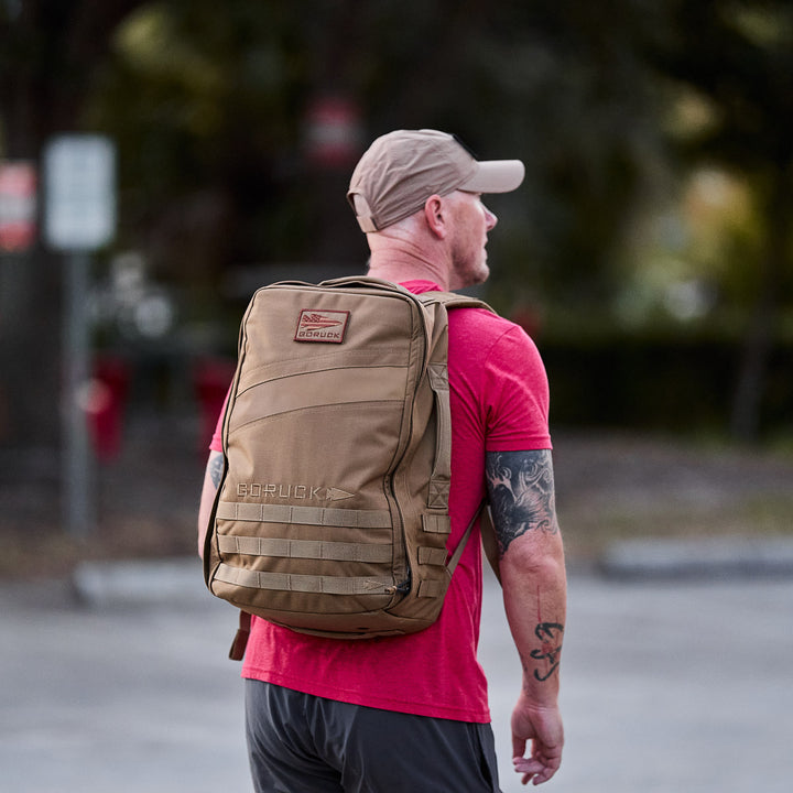 A man wearing a red shirt and cap stands outdoors in a parking lot, carrying the Rucker 4.0 tan rucking backpack.
