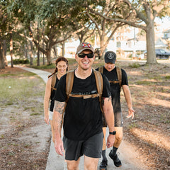 Three people wearing athletic clothes and Rucker 4.0 backpacks walk and smile on a tree-lined path in a park.