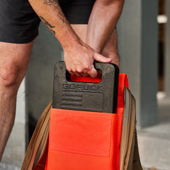 A person places a black GORUCK weight plate into the pocket of an orange Rucker 4.0 backpack, getting ready for a challenging workout.