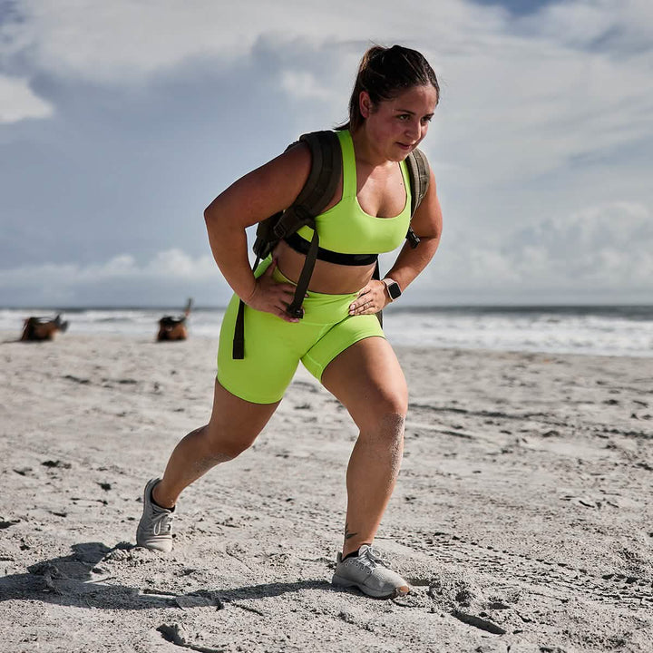 A person in a vibrant green athletic outfit, made with Toughflex Fabric by GORUCK, exercises on a sandy beach while wearing a weighted backpack. The ocean is visible under a partly cloudy sky and two dogs play near the shoreline in the distance.