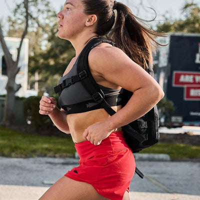 A woman sprints outdoors with a backpack, her Power Bra - Toughflex and red shorts moving seamlessly with each stride. Trees and vehicles dot the background as she enjoys the freedom of compression support.