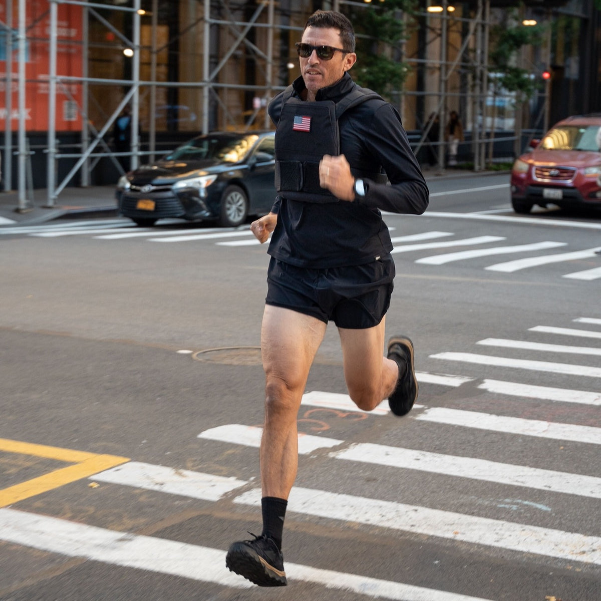 A man in black running gear jogs across a city street crosswalk in an urban setting, wearing the Rucking Weight Vest.