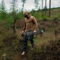 Man in outdoor GORUCK gear hiking in forest carrying compound bow and rugged backpack.