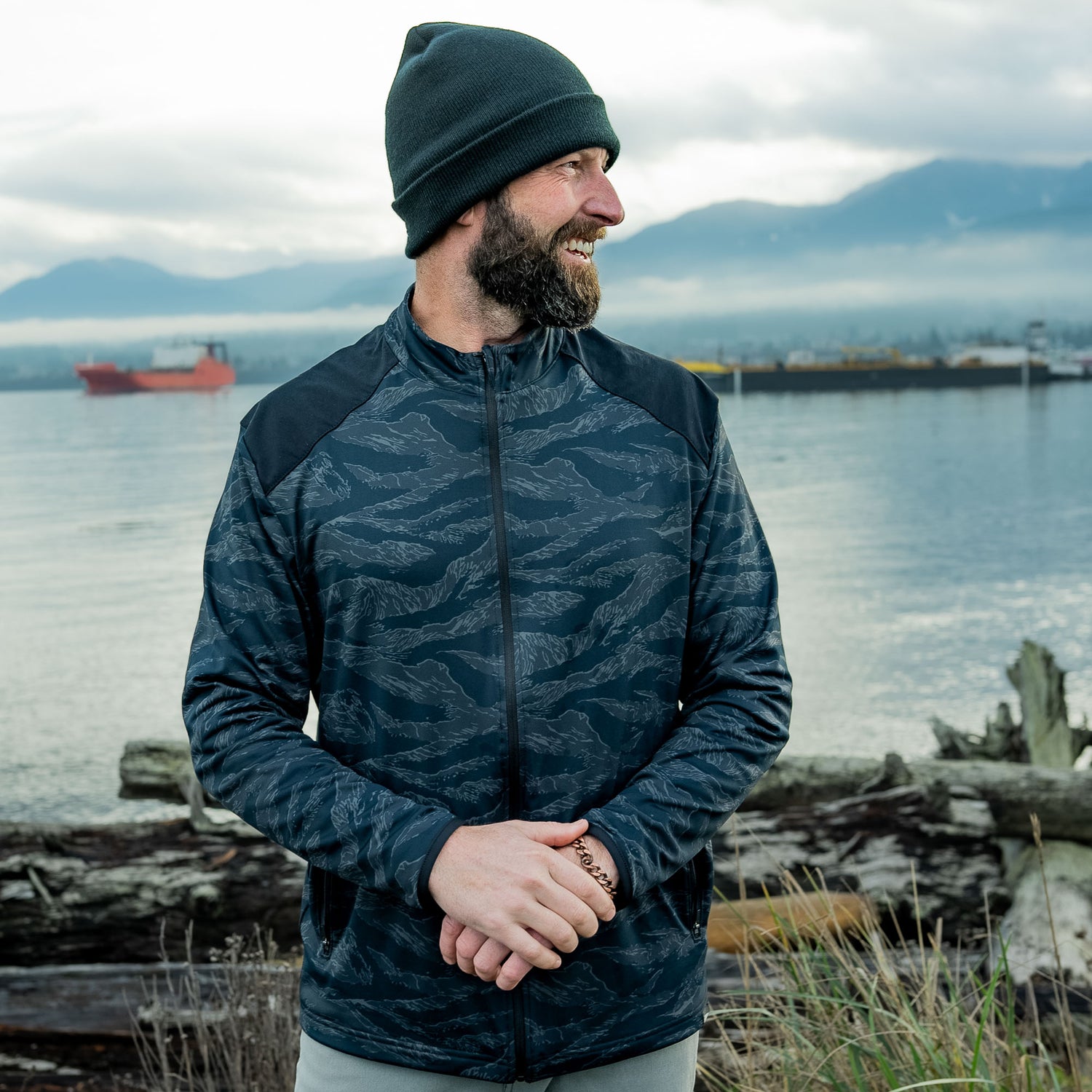 Man in GORUCK camo jacket and beanie outdoors by water, mountains and ship in background