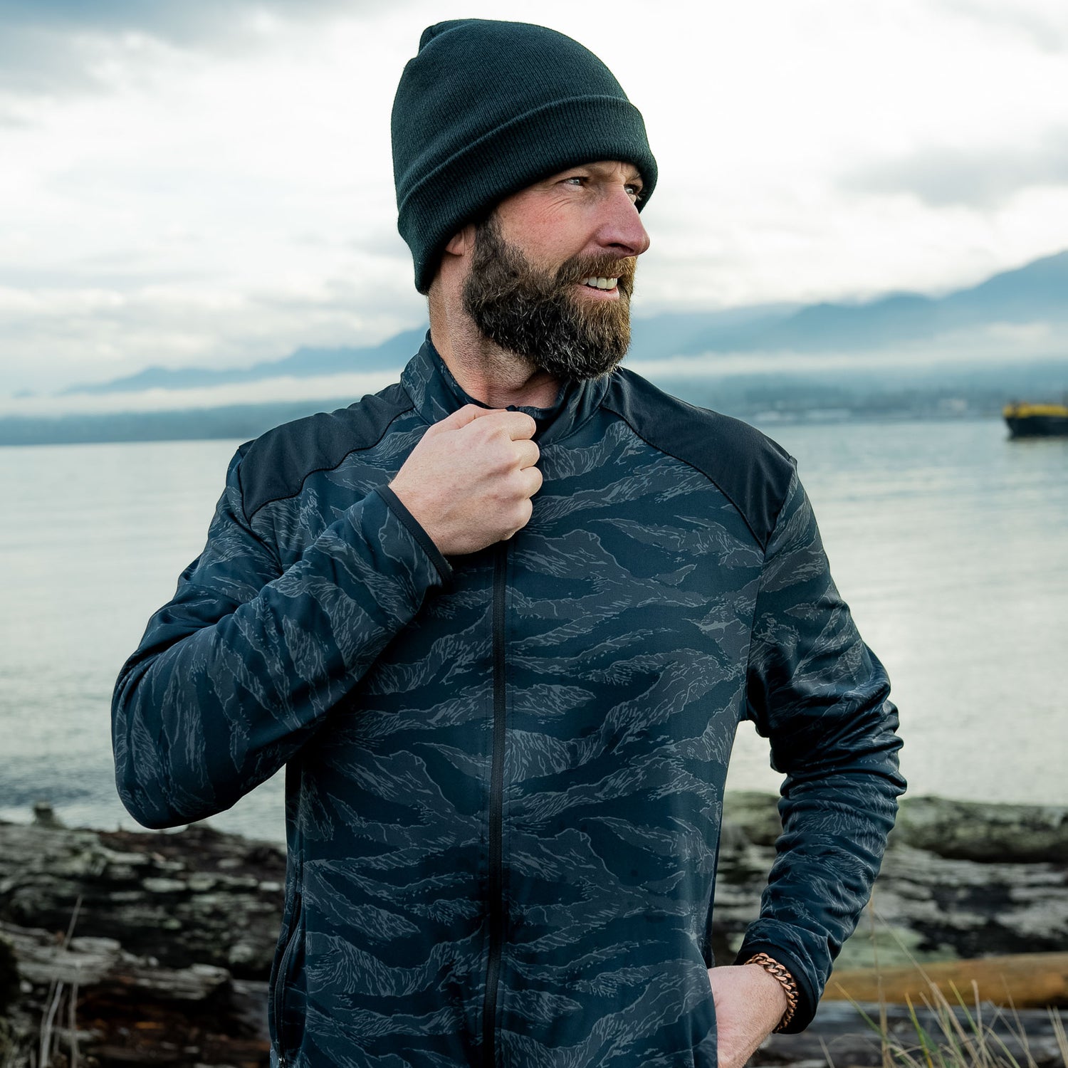 A bearded man in a beanie and patterned jacket, the Warm Up Full Zip, with zippered pockets, stands by the water, smiling and looking to the side.