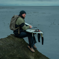 Man in beanie and rucking gear sitting on rock by water holding a surfboard with wetsuit