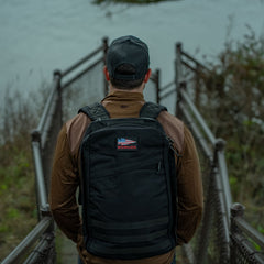 A person with a black GR1 USA - Cordura rucksack, recognized for its Special Forces grade durability, stands on a metal stairway overlooking a body of water.