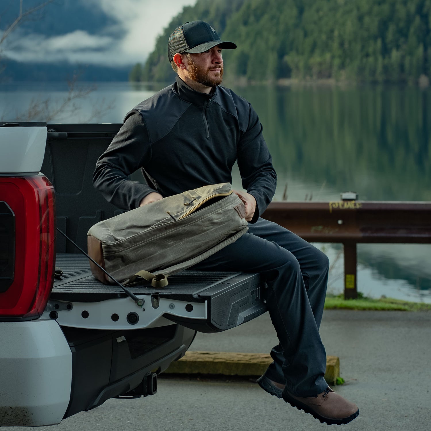 A man sits on a truck tailgate by a lake, holding a backpack and wearing outdoor clothing, including the Men’s Heavy Commando Quarter Zip - Merino Wool, known for odor resistance and moisture wicking on adventures.