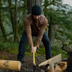 Man wearing a black beanie and brown long-sleeve shirt splitting wood with an axe outdoors in a forest
