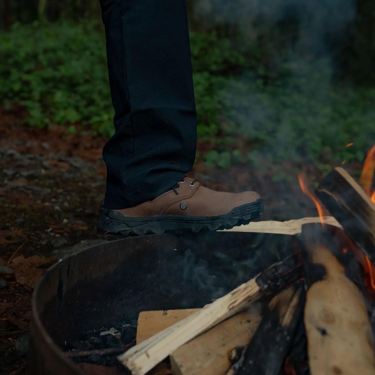 Close-up of rugged brown GORUCK boot stepping near a campfire with burning logs outdoors
