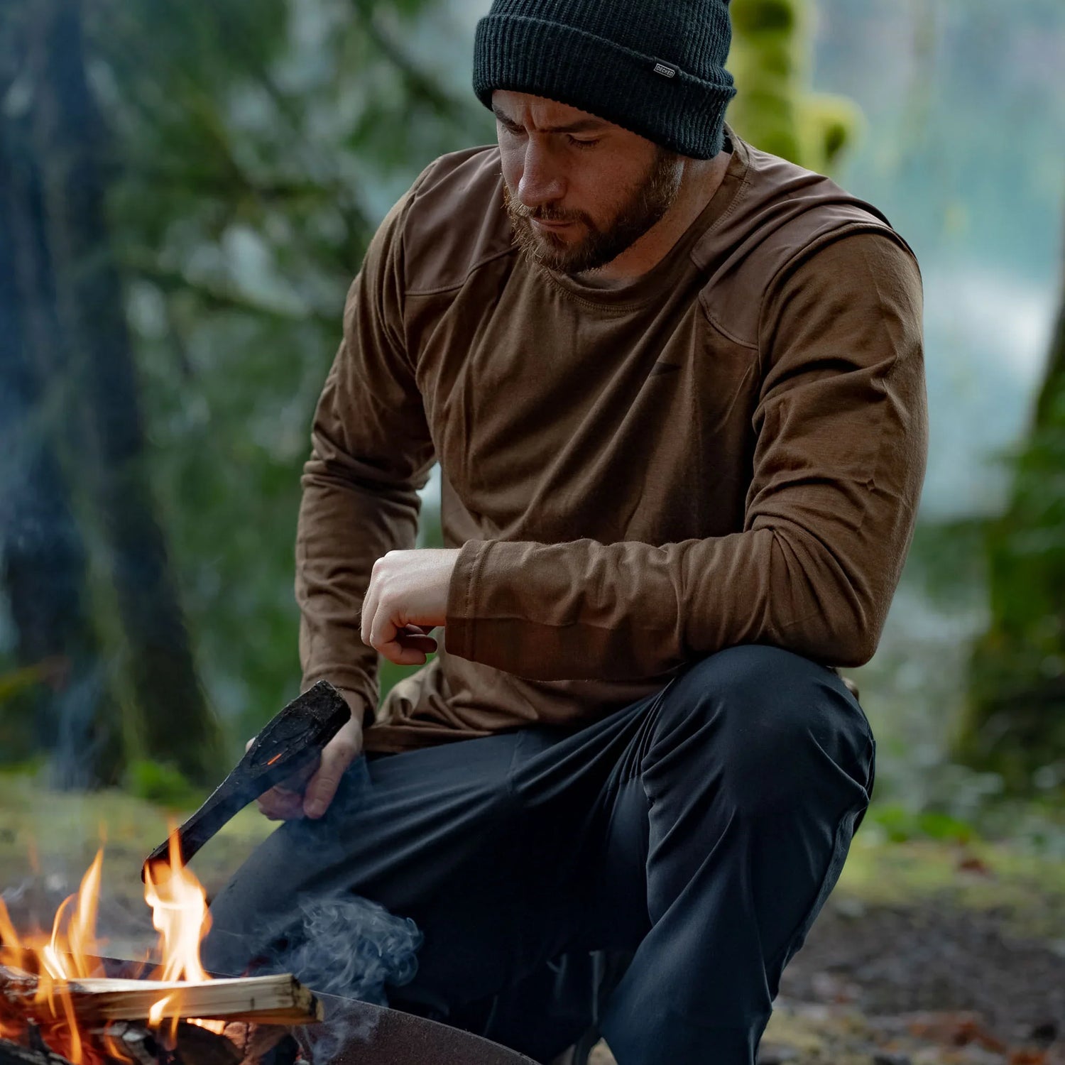 Man wearing beanie and brown long-sleeve shirt tending a campfire in outdoor forest setting