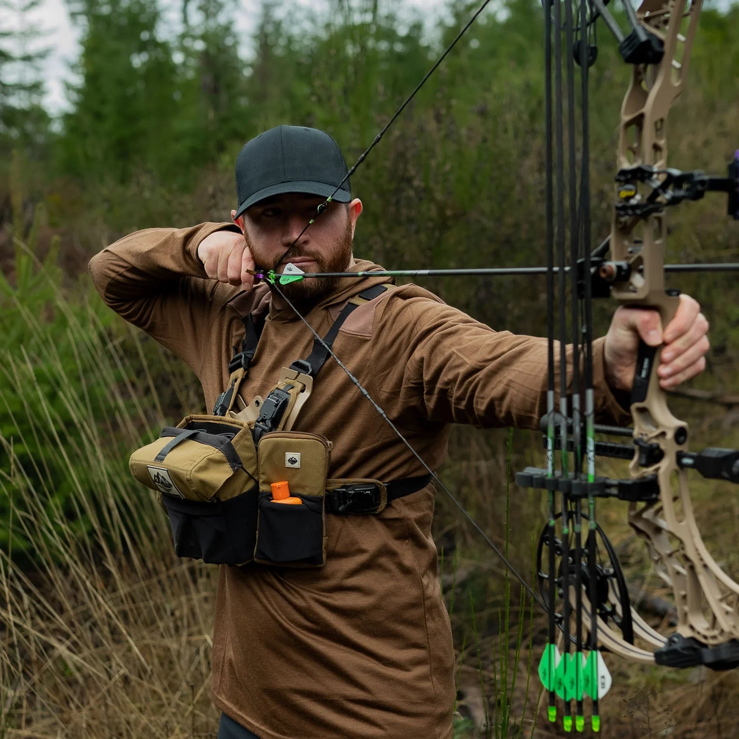 Man using compound bow with GORUCK chest pack outdoors in forest setting.